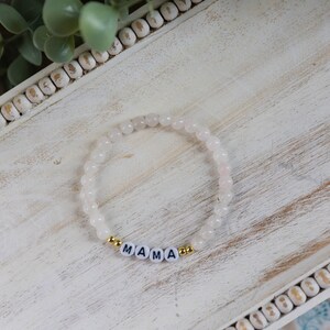 Könnte beinhalten: Weißes Perlenarmband mit goldenen Akzenten und dem Wort "MAMA" in weißen Buchstaben.