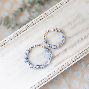 Könnte beinhalten: Zwei weiße Keramikperlenarmbänder mit blauen Blumendesigns. Ein Armband buchstabiert "MAMA" und das andere "MINI".