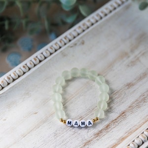 Könnte beinhalten: Ein hellgrünes Perlenarmband mit goldenen Abstandshaltern und weißen Buchstabenperlen, die "MAMA" buchstabieren.