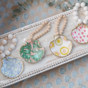 May include: Four decorative seashell ornaments with different patterns on a white tray. Each seashell is attached to a beaded garland with white beads. The tray has a white background with a beaded border.