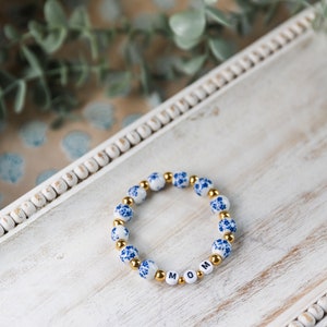 Könnte beinhalten: Ein Armband mit weißen Perlen mit blauen Blumenmustern und goldfarbenen Abstandshaltern. Das Armbandbuchstabiert "MOM" mit weißen Buchstabenperlen. Es wird auf einem weißen Holztablett präsentiert.