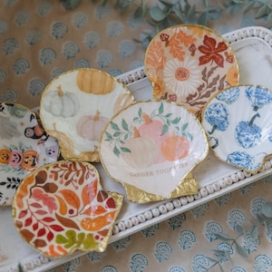 Puede incluir: Seis platos decorativos en forma de concha de vieira con ribete dorado y diversos diseños pintados. Los diseños incluyen calabazas, hojas de otoño, flores y un fantasma con una serpiente. Los platos están dispuestos en una bandeja blanca con un borde de cuentas.