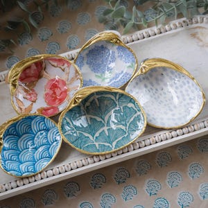 Op de afbeelding: Vijf decoratieve keramische schelpen met gouden rand. Elke schelp heeft een ander bloemen- of geometrisch patroon in tinten blauw, groen, roze en wit.