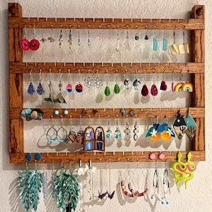 May include: A rectangular wooden earring organizer with four horizontal slats featuring evenly spaced notches. The rack holds a diverse collection of dangling earrings in various shapes, colors, and designs, including floral, geometric, and animal motifs.