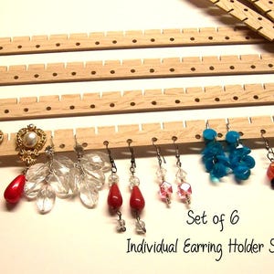 May include: Set of six individual wooden earring holder slats. The slats are light brown and feature evenly spaced holes for displaying earrings. Various earrings with red, clear, and blue accents are displayed.