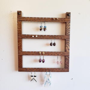 May include: A rectangular dark brown wooden earring organizer featuring four horizontal slats with pre-drilled holes for hanging jewelry. The frame displays a visible wood grain texture and holds various pairs of dangling and stud earrings.