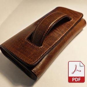 Puede incluir: Un estuche rectangular de cuero marrón con un asa superior y cierre de solapa. El cuero tiene una textura visible y costuras tonales a lo largo de los bordes. El estuche está orientado en diagonal, mostrando el asa y la superficie oscura.