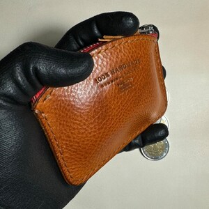 May include: A brown leather coin purse with a red zipper and the text "100% HANDMADE" embossed on the front.