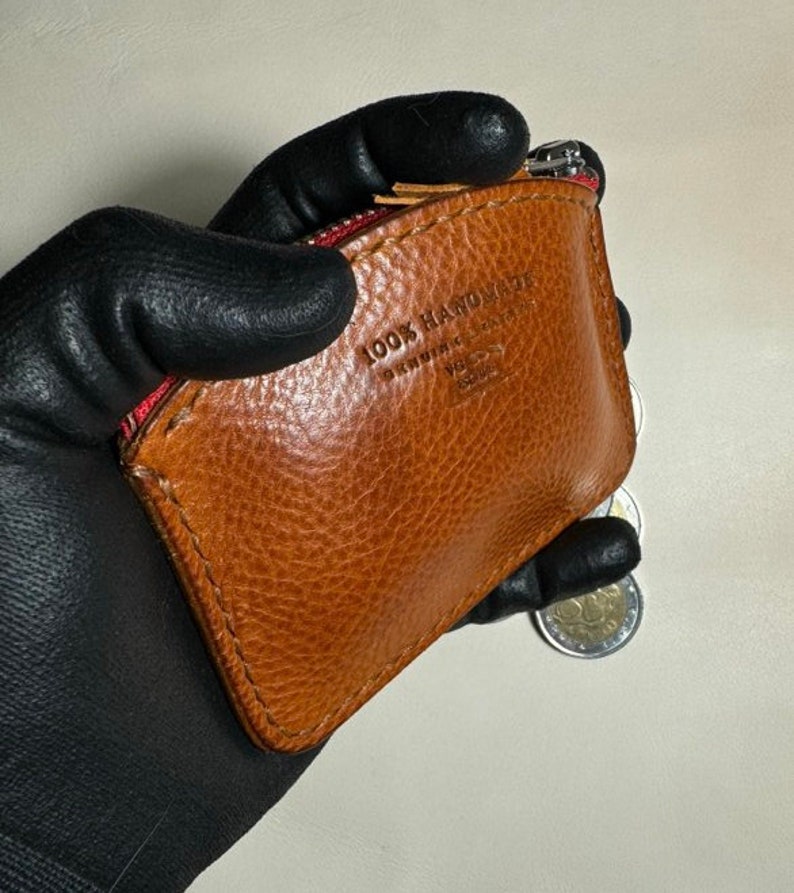 May include: A brown leather coin purse with a red zipper and the text "100% HANDMADE" embossed on the front.