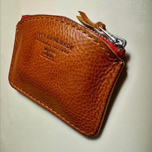 May include: A brown leather coin purse with a red zipper and the text "100% HANDMADE GENUINE LEATHER WE LOVE  EZRA" embossed on the front.