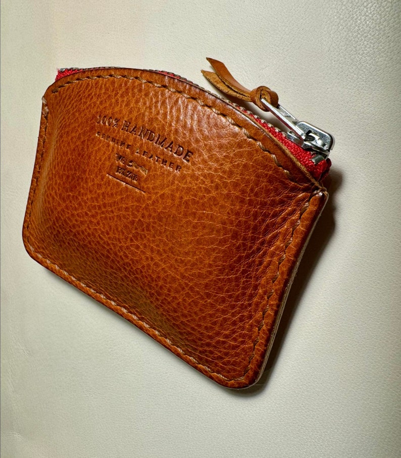 May include: A brown leather coin purse with a red zipper and the text "100% HANDMADE GENUINE LEATHER WE LOVE  EZRA" embossed on the front.