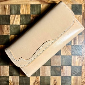 May include: A light tan leather wallet with a curved flap and white stitching. The wallet is rectangular and appears to be closed. The background is a wooden checkerboard pattern.