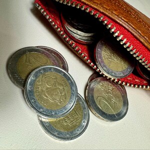 May include: A brown leather coin purse with a red zipper is open, revealing a collection of euro coins. The coins are silver and gold in color and feature various designs.