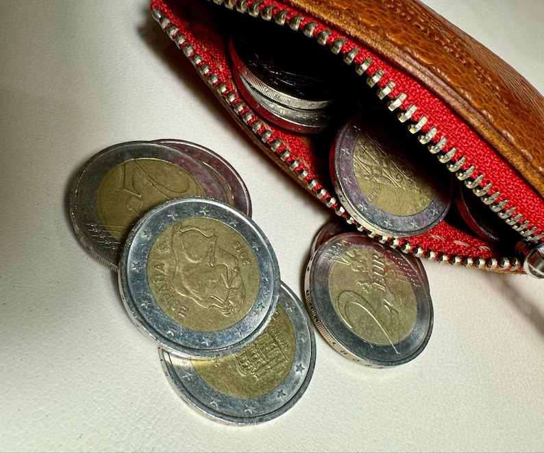 May include: A brown leather coin purse with a red zipper is open, revealing a collection of euro coins. The coins are silver and gold in color and feature various designs.