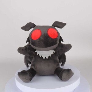 May include: A plush toy shaped like a moth with large red eyes and a white furry collar. The moth is sitting on a white platform.