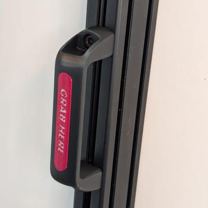 Puede incluir: Un asa negra con una etiqueta roja que dice "GRAB HERE" está fijada a un marco de metal negro. El asa está diseñada para un agarre fácil y probablemente forma parte de una estructura o mecanismo más grande.