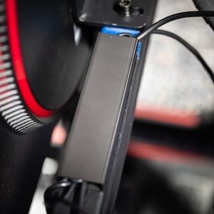 以下が含まれることがあります： 黒色の金属製のブラケットのクローズアップ。背景には赤と白の歯車があります。ブラケットには青いワイヤーが取り付けられています。