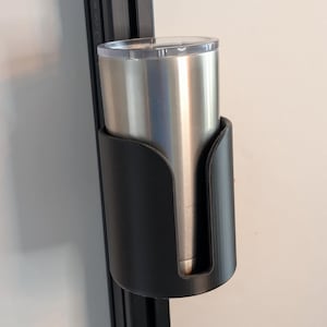 May include: A black plastic cup holder mounted on a black metal track. A silver stainless steel travel mug with a clear lid is in the holder.
