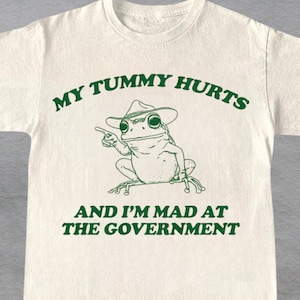 May include: A white t-shirt featuring a green cartoon frog wearing a hat and the text "My tummy hurts and I'm mad at the government".