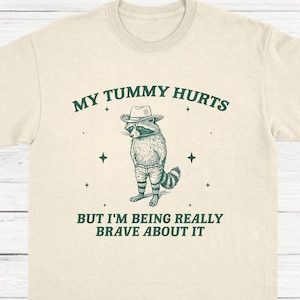 May include: Cream-coloured t-shirt featuring a raccoon illustration in a hat and shorts. The text "MY TUMMY HURTS BUT I'M BEING REALLY BRAVE ABOUT IT" is printed above and below the raccoon graphic. The shirt has a crew neck and short sleeves.