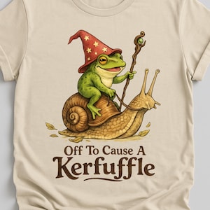 Puede incluir: Camiseta beige con una ilustración de una rana verde con sombrero de mago montada en un caracol. La rana sostiene un bastón y el texto "Off To Cause A Kerfuffle" está debajo de la imagen.