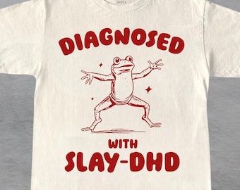 Può includere: Maglietta bianca con una grafica rossa di una rana con il testo "Diagnosed with Slay-DHD".