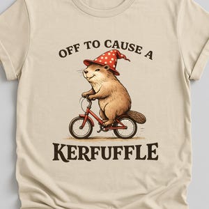 Könnte beinhalten: Beiges T-Shirt mit einer Grafik eines braunen Nagetiers mit rotem Hut und weißen Punkten, das ein rotes Fahrrad fährt. Der Text oben lautet "OFF TO CAUSE A" und darunter "KERFUFFLE".