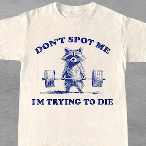 May include: White t-shirt with a blue graphic of a raccoon lifting a barbell. The text "Don't Spot Me I'm Trying To Die" is printed below the raccoon.