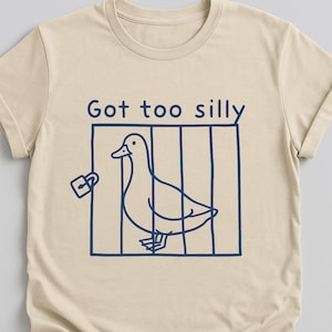 以下が含まれることがあります： ベージュのTシャツに、青い鳥かごの中の鴨のグラフィックが描かれています。グラフィックの上に「Got too silly」というテキストがあります。