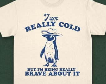 Puede incluir: Una camiseta blanca con un pingüino azul que lleva un sombrero de vaquero. El texto de la camiseta dice "Tengo mucho frío, pero soy muy valiente al respecto."