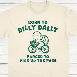 May include: A beige t-shirt with a green graphic of a turtle riding a bicycle. The text "Born to Dilly Dally" and "Forced to Pick Up the Pace" is printed on the shirt.