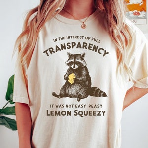 May include: A beige t-shirt with a graphic of a raccoon holding a lemon. The text on the shirt reads "In the interest of full transparency, it was not easy peasy lemon squeezy."
