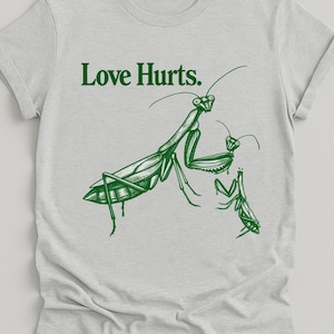 May include: A light gray short-sleeve t-shirt featuring a dark green graphic of two praying mantises and the text "Love Hurts." printed above the insects. The design is centered on the chest, showcasing detailed line work of the mantises.