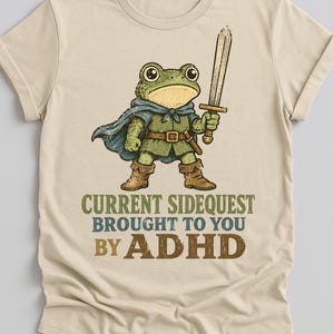 May include: A beige short-sleeved t-shirt featuring a graphic of a green frog dressed as a warrior with a blue cape, brown boots, and a sword. Below the frog, the text reads: CURRENT SIDEQUEST BROUGHT TO YOU BY ADHD in green, blue, and brown.