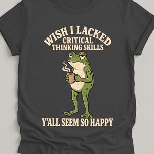 Op de afbeelding: Een antracietgrijs t-shirt met een afbeelding van een groene kikker die een dampende bruine mok vasthoudt. De tekst boven de kikker luidt WISH I LACKED CRITICAL THINKING SKILLS en de tekst eronder luidt Y'ALL SEEM SO HAPPY in crèmekleur.
