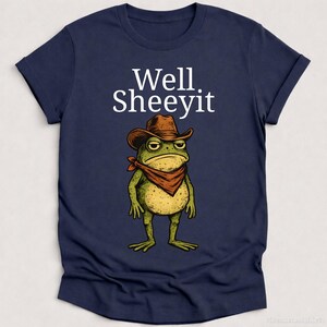 K&ouml;nnte beinhalten: Marineblaues T-Shirt mit einer Cartoon-Fr&ouml;schin, die einen Cowboyhut und ein rotes Halstuch tr&auml;gt. Der Text "Well Sheeyit" ist &uuml;ber dem Frosch aufgedruckt. Der Frosch hat einen m&uuml;rrischen Gesichtsausdruck.