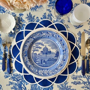 Op de afbeelding: Een tafelsetting met een blauw en wit toile de Jouy-patroon. Inclusief een blauw bord met een landschapssc&egrave;ne, een blauwe placemat, zilverwerk met blauwe handvatten en een wit blad-vormig schaaltje. Een blauw glas en een wit glas zijn ook aanwezig.