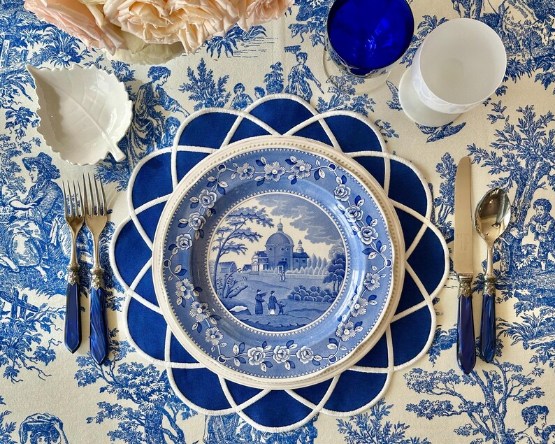 Op de afbeelding: Een tafelsetting met een blauw en wit toile de Jouy-patroon. Inclusief een blauw bord met een landschapssc&egrave;ne, een blauwe placemat, zilverwerk met blauwe handvatten en een wit blad-vormig schaaltje. Een blauw glas en een wit glas zijn ook aanwezig.