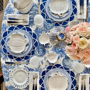 Op de afbeelding: Een tafelsetting met witte pompoenvormige borden, blauw-witte geruite borden en blauwe bestekken. De tafel is bedekt met een blauw-wit toile de jouy tafelkleed, roze rozen en blauw glaswerk.