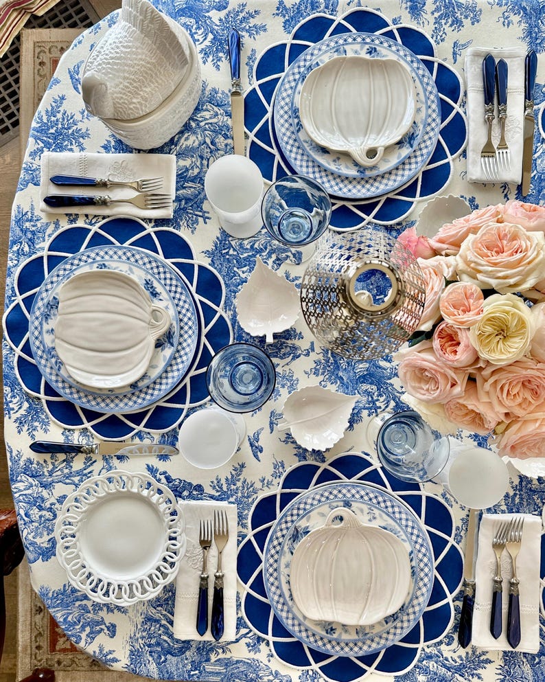 Op de afbeelding: Een tafelsetting met witte pompoenvormige borden, blauw-witte geruite borden en blauwe bestekken. De tafel is bedekt met een blauw-wit toile de jouy tafelkleed, roze rozen en blauw glaswerk.