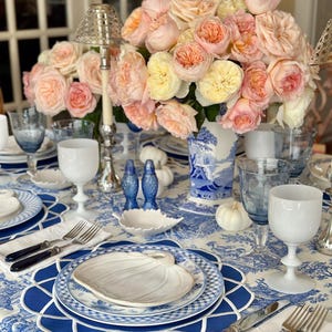 Op de afbeelding: Een prachtig gedekte tafel met een blauw-wit bloemig tafelkleed, met een middelpunt van perzik- en gele rozen in een blauw-witte vaas. Inclusief wit en blauw glaswerk, borden en bestek.