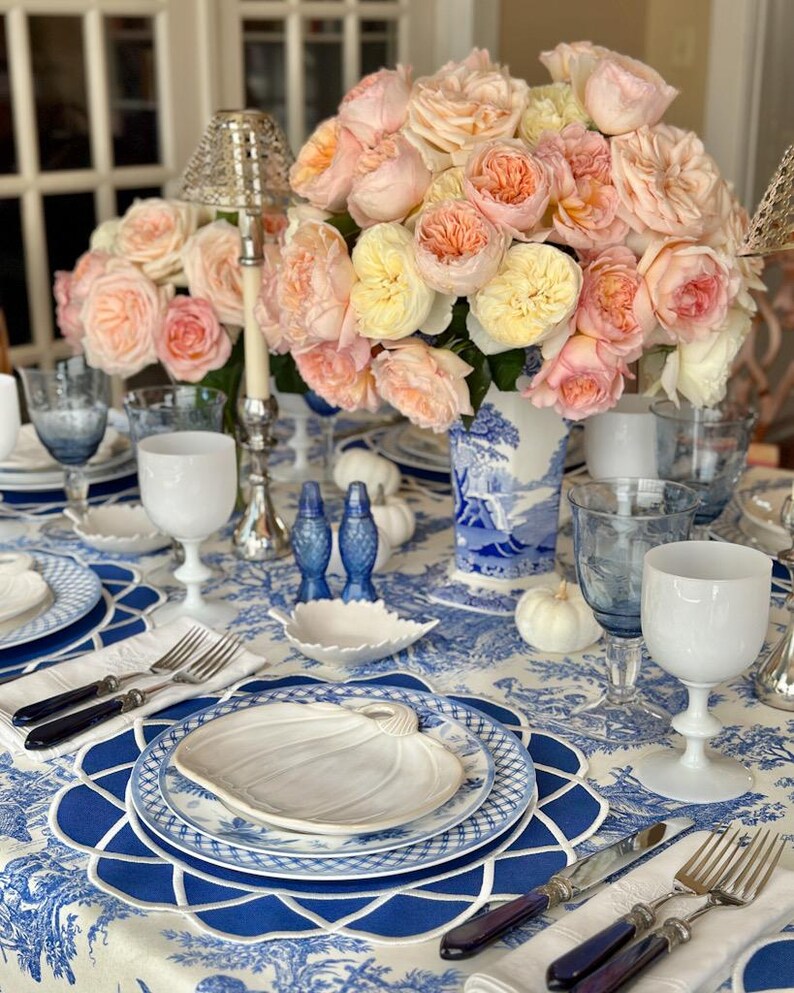 Op de afbeelding: Een prachtig gedekte tafel met een blauw-wit bloemig tafelkleed, met een middelpunt van perzik- en gele rozen in een blauw-witte vaas. Inclusief wit en blauw glaswerk, borden en bestek.