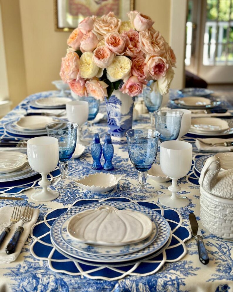 Op de afbeelding: Elegante tafelsetting met een blauw-wit toile de Jouy tafelkleed. Voorzien van wit en blauw glaswerk, borden en een centerpiece van roze en gele rozen in een blauw-witte vaas. Inclusief een wit pompoenvormig bord.