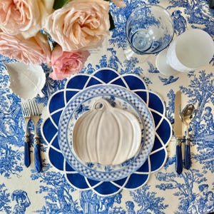 Op de afbeelding: Een tafelsetting met een blauw-wit toile de jouy tafelkleed, met een witte pompoenvormige bord, blauwe borden en een blauwe geschulpte placemat. Zilverwerk met blauwe handvatten en roze rozen maken de elegante opstelling compleet.