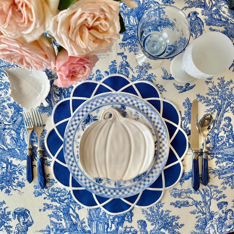 Op de afbeelding: Een tafelsetting met een blauw-wit toile de jouy tafelkleed, met een witte pompoenvormige bord, blauwe borden en een blauwe geschulpte placemat. Zilverwerk met blauwe handvatten en roze rozen maken de elegante opstelling compleet.
