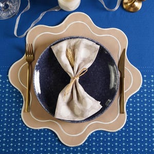 May include: A beige placemat with a scalloped edge and a white border surrounds a dark blue ceramic plate with a white linen napkin folded on top. A wheat stalk is tied with twine and placed on top of the napkin. A gold fork and knife are placed on the placemat.