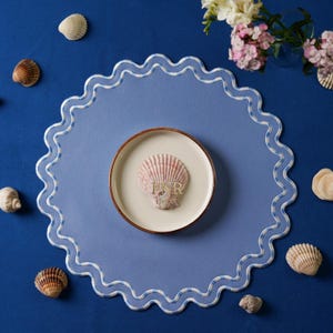 May include: A blue placemat with a white scalloped edge and a white and blue checkered pattern. A small white plate with a pink seashell on top is in the center of the placemat. The placemat is on a blue surface with several seashells scattered around it.