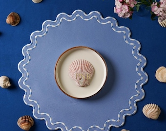 May include: A blue placemat with a white scalloped edge and a white and blue checkered pattern. A small white plate with a pink seashell on top is in the center of the placemat. The placemat is on a blue surface with several seashells scattered around it.