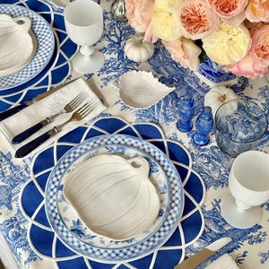Op de afbeelding: Een prachtig gedekte tafel met een blauw-wit toile de Jouy tafelkleed. De tafelsetting omvat witte pompoenvormige borden, blauwe borden, witte wijnglazen en zilverwerk. Een boeket roze en gele rozen.