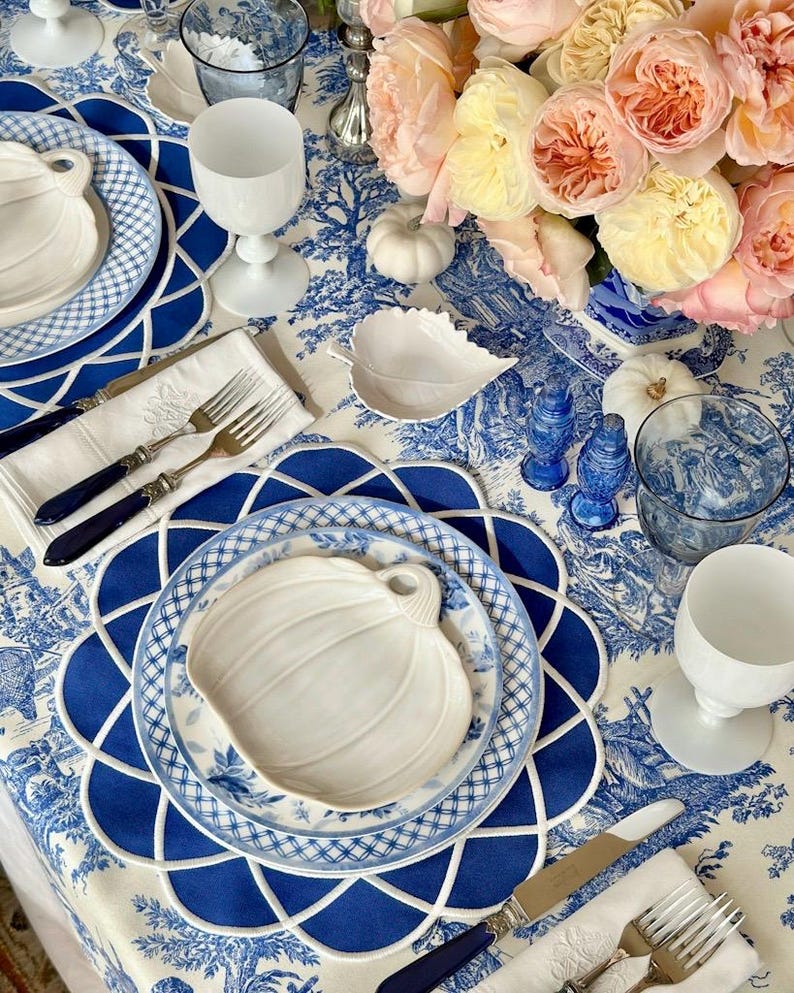 Op de afbeelding: Een prachtig gedekte tafel met een blauw-wit toile de Jouy tafelkleed. De tafelsetting omvat witte pompoenvormige borden, blauwe borden, witte wijnglazen en zilverwerk. Een boeket roze en gele rozen.